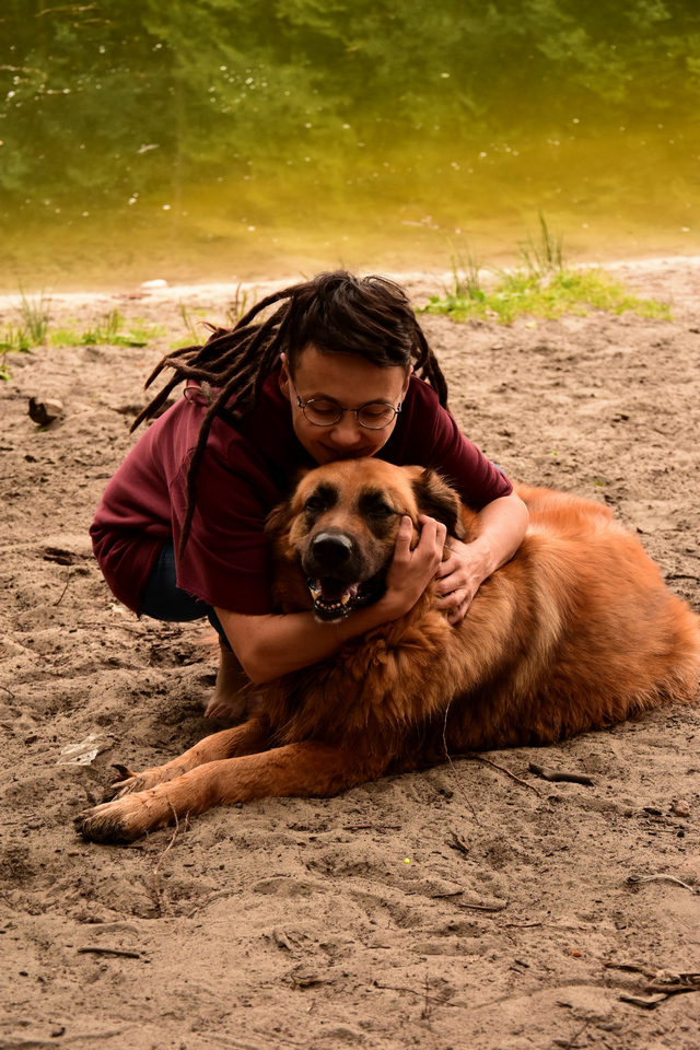 dziewczyna w dredach obejmująca rudego, wielkiego psa na tle jeziora w lesie